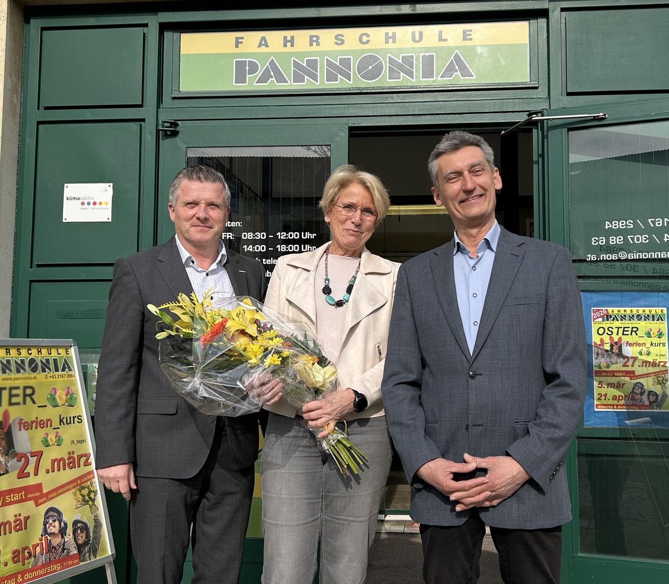 Drei Personen, zwei Männer und eine Frau, stehen vor einem Gebäude. Sie lächeln und halten Blumensträuße in den Händen. Die Frau trägt eine Halskette, und die Männer sind in Anzügen gekleidet. Hinter ihnen befindet sich ein Schild mit der Aufschrift 'FAHRSCHULE PANNONIA'.