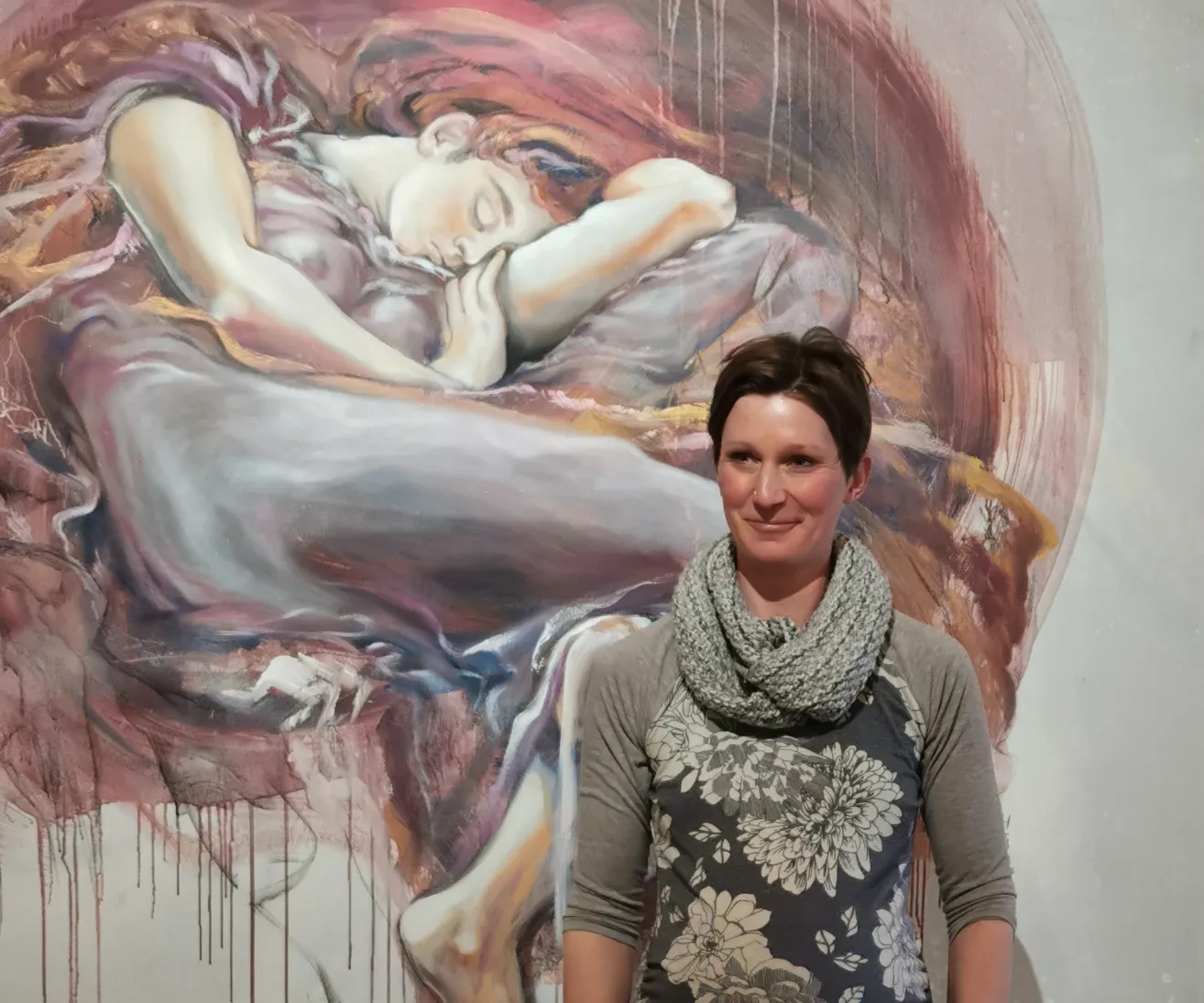 Eine Frau steht vor einem großen Gemälde und lächelt für ein Foto. Das Gemälde zeigt eine schlafende Frau, möglicherweise ein Porträt. Das Gemälde hat einen einzigartigen Stil mit Farbspritzern und abstrakten Elementen.
