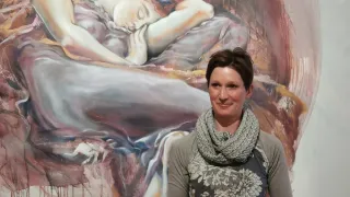 Eine Frau steht vor einem großen Gemälde und lächelt für ein Foto. Das Gemälde zeigt eine schlafende Frau, möglicherweise ein Porträt. Das Gemälde hat einen einzigartigen Stil mit Farbspritzern und abstrakten Elementen.