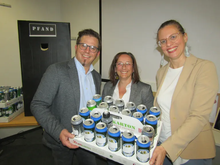 Three people, two women and one man, are holding a box filled with cans of beer. They are smiling and appear to be posing for a photo.