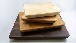 Three wooden cutting boards stacked on top of each other, one with a darker finish and two lighter ones, arranged on a white surface.
