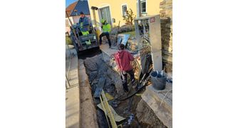 A few workers are digging the ground to lay cables in front of a house. The man in the back is operating a construction vehicle. The others are working with tools and cables.