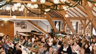 Eine große Gruppe von Frauen sitzt an Tischen in einem festlich geschmückten Saal mit Holzbalken, verziert mit Grünpflanzen und hängenden Lampen, was auf eine gesellschaftliche Zusammenkunft oder Veranstaltung hindeutet.