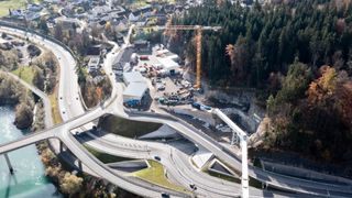 An aerial view of a road construction site with a bridge and a crane. Buildings, cars, and construction vehicles are scattered around the area.