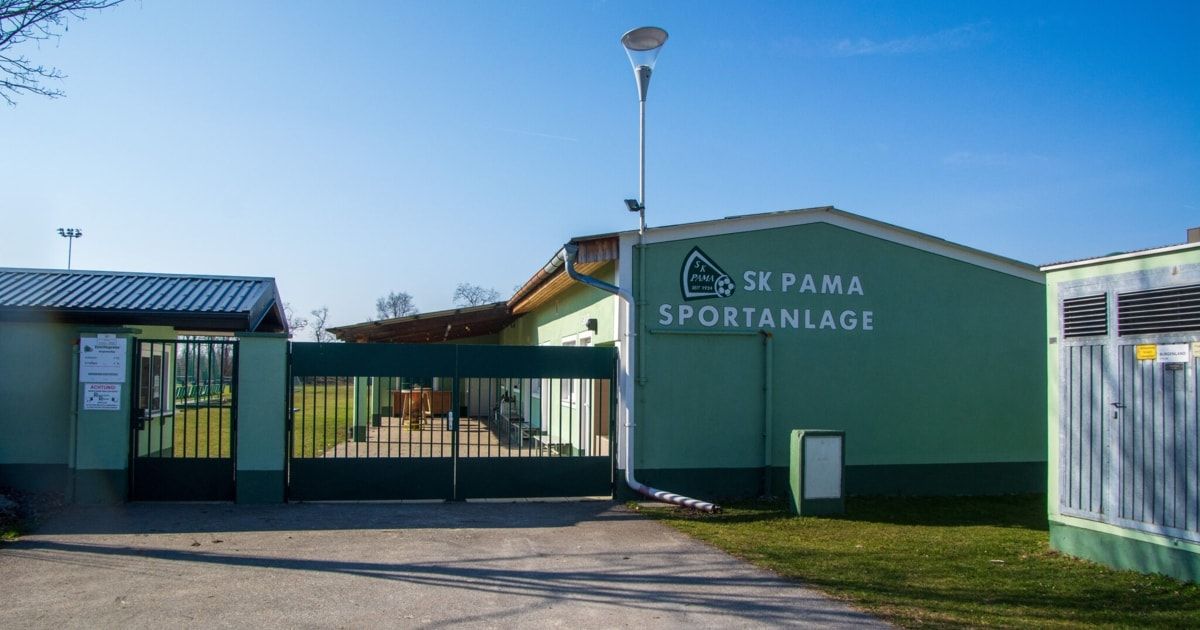 Die SK PAMA-Sportanlage, ein grünes Gebäude mit einem Tor, hat ein Logo an der Seite und eine Lampe über dem Tor.