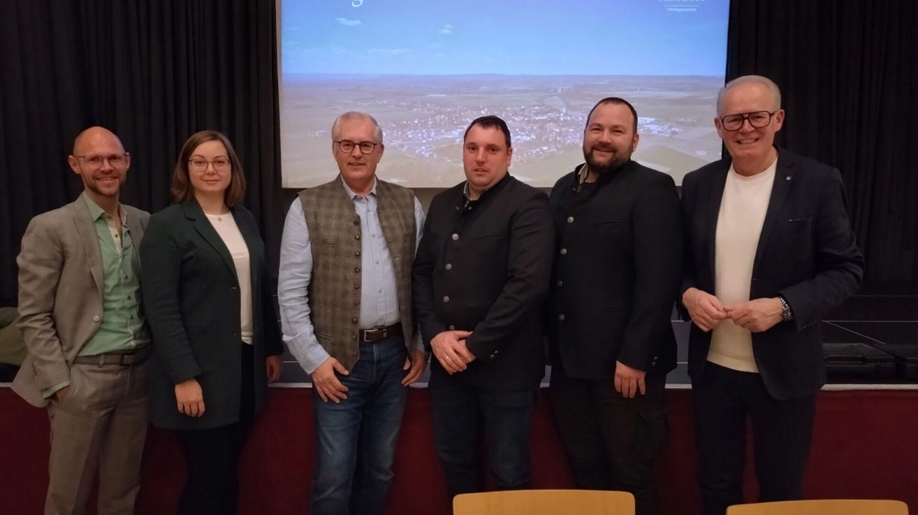 Five people stand before a screen showing a landscape, dressed formally. They appear to be posing for a photograph.