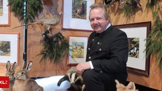 A man in a black uniform sits next to a badger and a fox, with wildlife paintings on the wooden wall behind him.