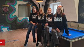 Vier Personen in schwarzen Hoodies mit den Worten 'SpieK TRUM' sitzen lächelnd auf einem Billardtisch. Sie befinden sich in einem Raum mit einer bunten Wand und einem Lautsprecher.