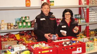 Zwei Personen stehen in einem Lagerraum, halten Lebensmittelboxen in der Hand und lächeln. Hinter ihnen befinden sich Regale mit verschiedenen Produkten. Die Person auf der linken Seite hält eine Box mit Snacks und die auf der rechten Seite eine Box mit Süßigkeiten.