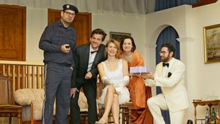 Eine Gruppe von Menschen in Vintage-Kleidung, lächelnd, posiert für ein Foto. Die Frau in der Mitte hält ein Geschenk.