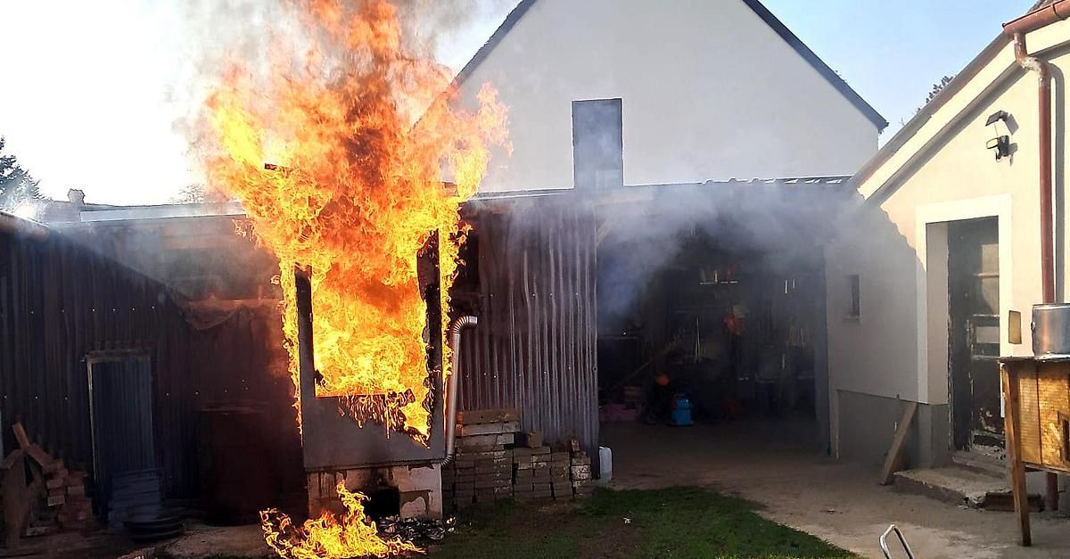 Ein Holzofen ist von Flammen und Rauch umhüllt und steht vor einer Garage mit einer Metallwand.