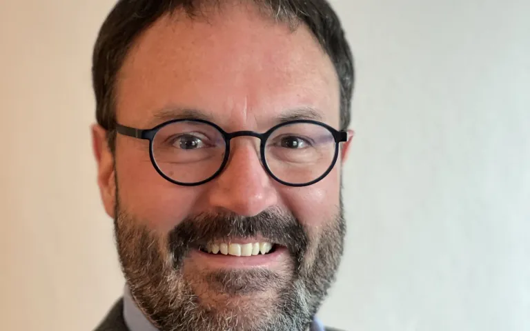 Ein Mann mit Brille und Bart lächelt in die Kamera und trägt ein graues Hemd.