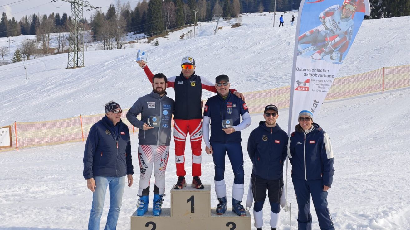 A group of men stands on a snowy podium with awards, two wearing red and white ski suits, one in a black jacket with a logo, and others in blue jackets.
