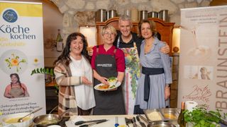 Vier Menschen stehen lächelnd in einer Küche. Einer hält einen Teller mit Essen. Zwei tragen Schürzen. Der Hintergrund zeigt Steinwände und Regale mit Flaschen.