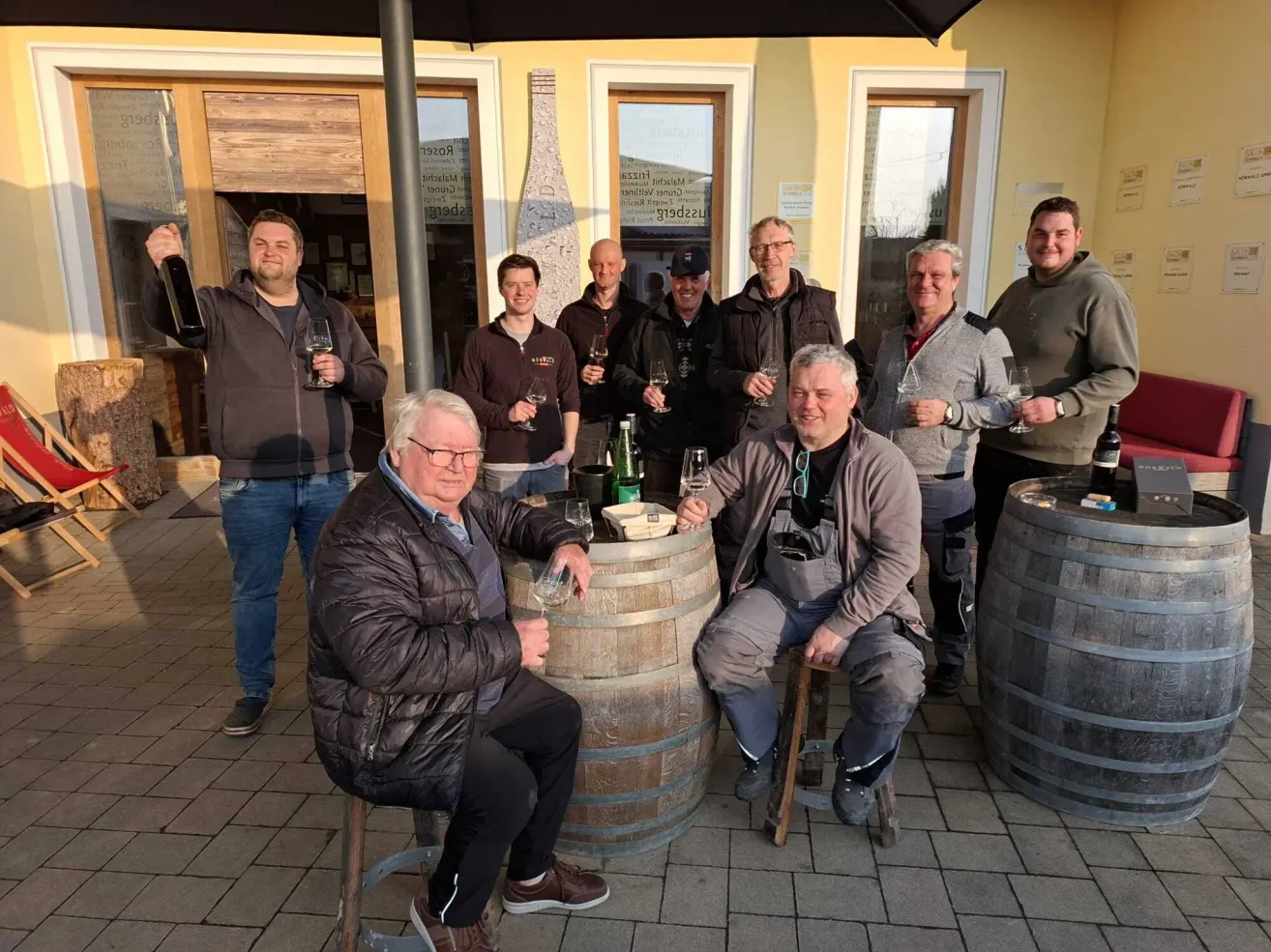 A group of men sit around a barrel, posing for a photo. Some hold wine glasses, and a man holds a bottle. Behind them, a building with glass doors and windows is visible.