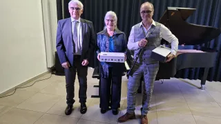Three individuals, two men and a woman, stand in a room with a piano in the background. They are all smiling and holding boxes. The room has tiled flooring and a black curtain.