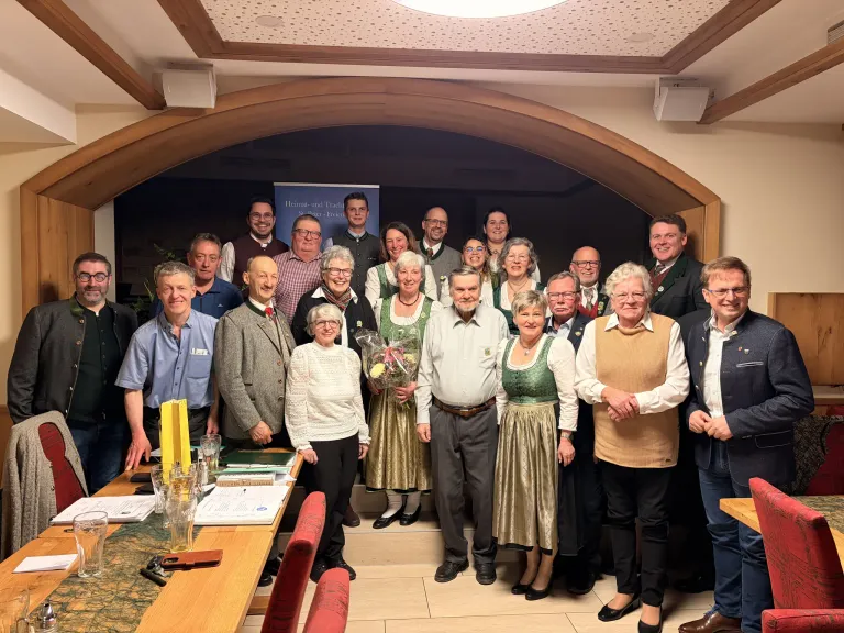Eine Gruppe von Menschen in traditioneller Kleidung, die lächelnd und für ein Foto in einem Bankettsaal mit Holztischen, einem dekorativen Bogen und einem Banner im Hintergrund posieren.