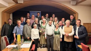 Eine Gruppe von Menschen in traditioneller Kleidung, die lächelnd und für ein Foto in einem Bankettsaal mit Holztischen, einem dekorativen Bogen und einem Banner im Hintergrund posieren.