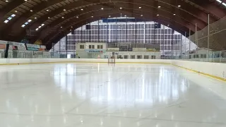 An empty indoor ice hockey rink with a high arched ceiling, yellow border, and large windows. Several lights are mounted on the ceiling.