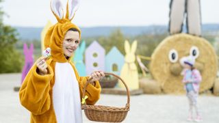 A woman dressed in a rabbit costume, holding a basket, stands in front of a backdrop with colorful houses and a large haystack.