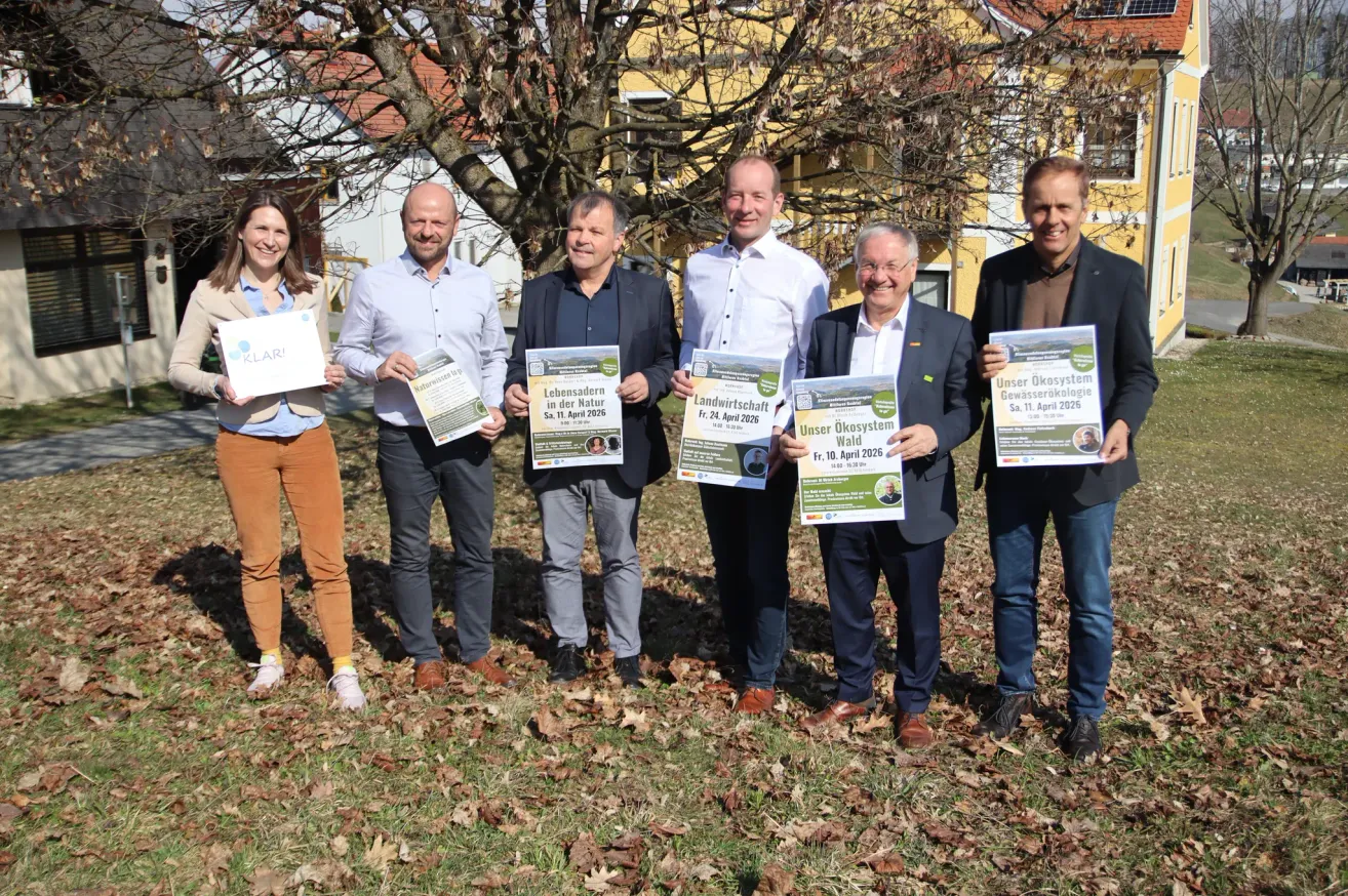Five people are standing in a field, smiling. They hold signs that mention events and dates. Behind them is a house with yellow walls and a tree.