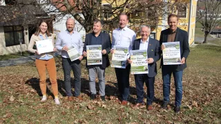 Five people are standing in a field, smiling. They hold signs that mention events and dates. Behind them is a house with yellow walls and a tree.