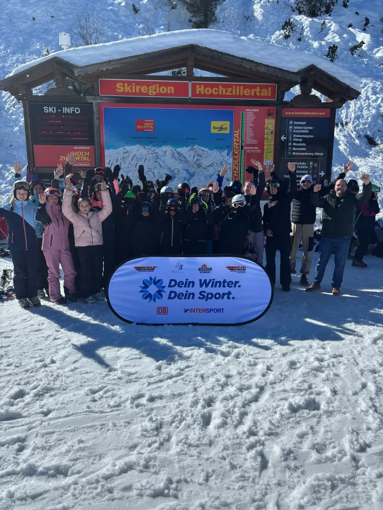 Eine Gruppe von Menschen, die Winterkleidung tragen, steht in einem verschneiten Bereich vor einem Schild, das 'Skiregion Hochzillertal' sagt. Sie alle lächeln und scheinen für ein Foto zu posieren.
