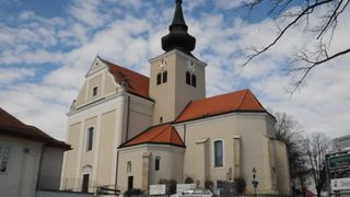 A beige church with red roofs and a tall steeple. The steeple has two clocks and is topped with a black onion dome.
