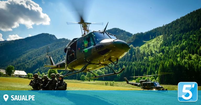 Military helicopter in flight with soldiers on ground. Surrounded by a mountainous landscape with lush green fields and trees.