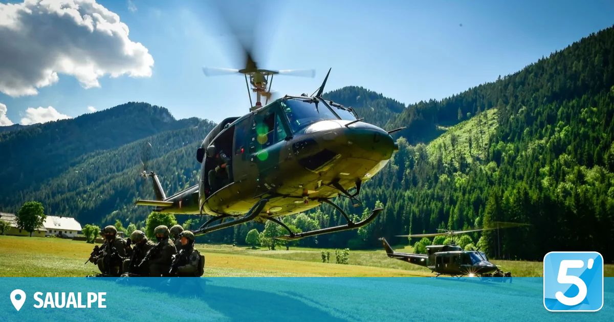 Military helicopter in flight with soldiers on ground. Surrounded by a mountainous landscape with lush green fields and trees.