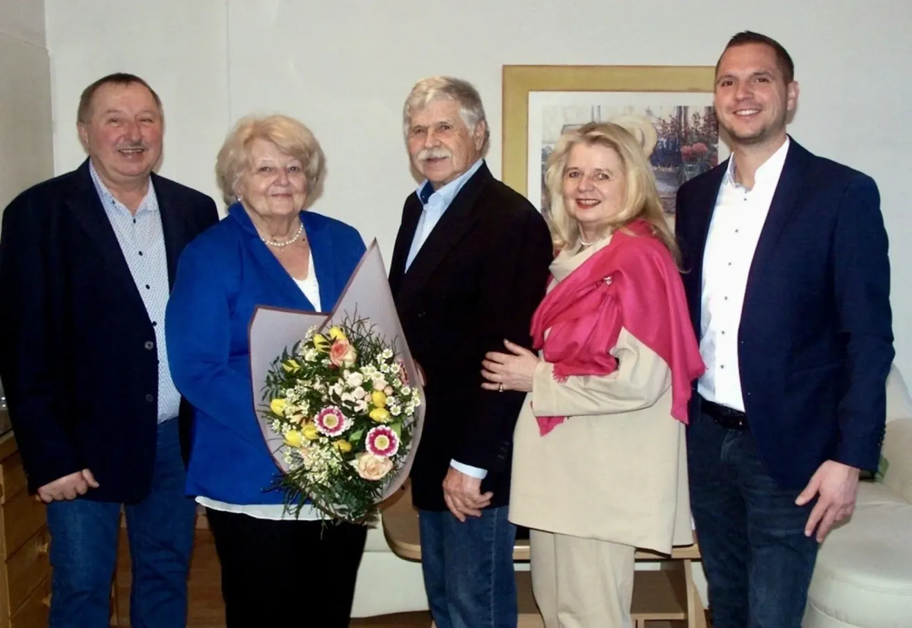 Fünf Personen, darunter zwei Frauen mit Blumen, stehen für ein Foto in einem Raum. Ein gerahmtes Bild hängt an der Wand hinter ihnen.