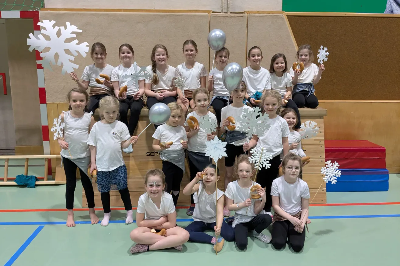 A group of girls in white shirts and pants, some wearing glasses, are posing for a photo while holding white snowflake-shaped objects and balloons. They are seated on the floor and on a bench, with a wooden wall in the background.
