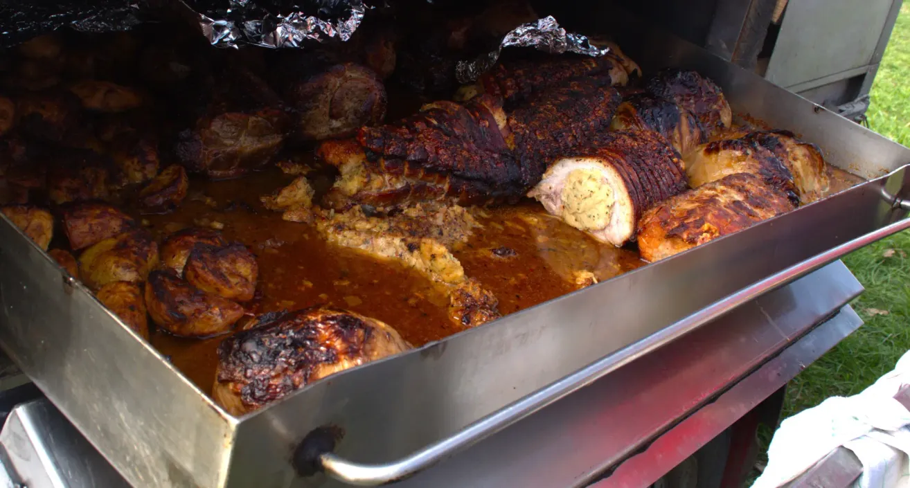 A close-up view of various meats being cooked in a smoker, covered in foil, and basted with a red sauce.