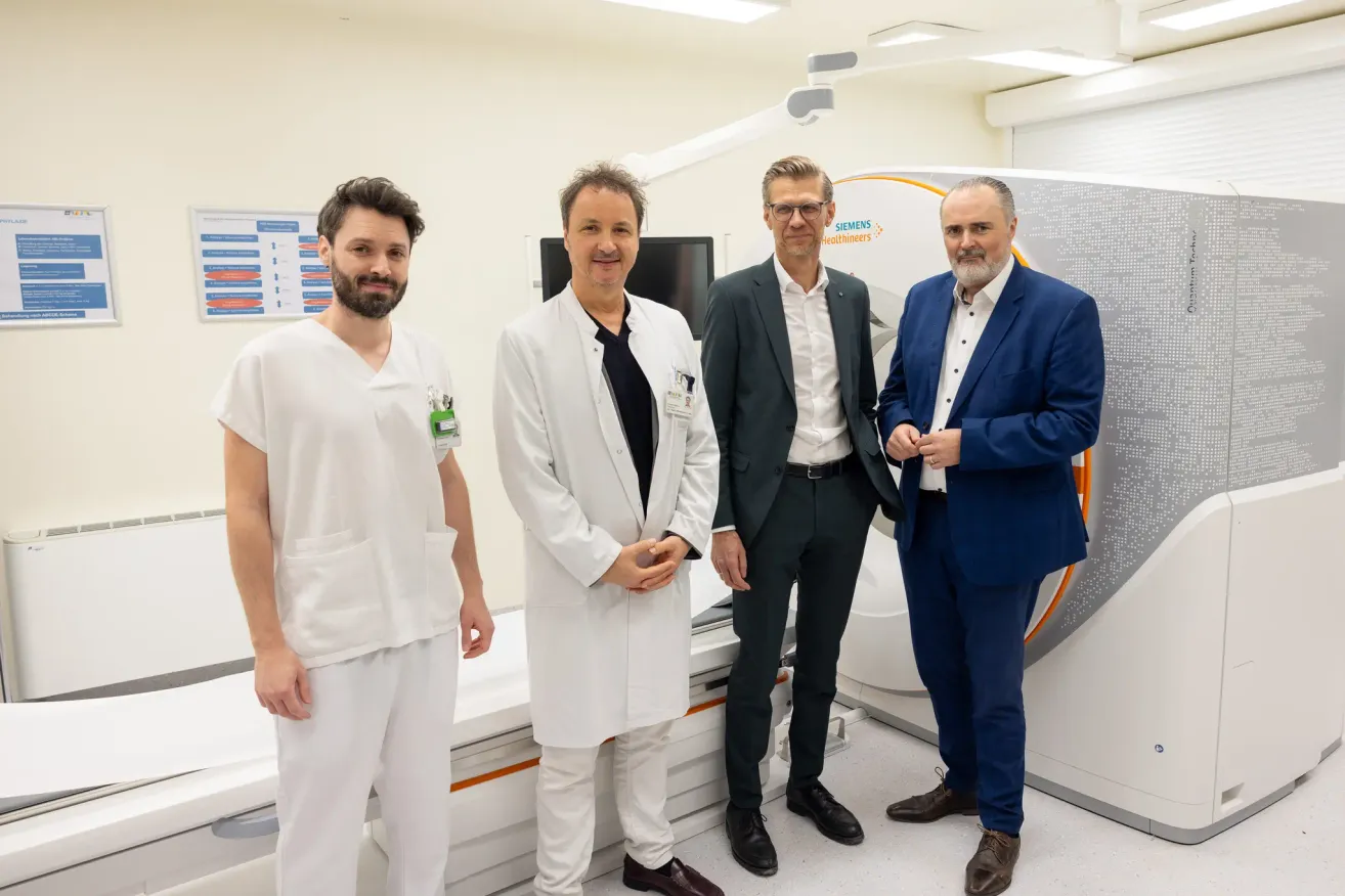 Vier Männer stehen in einem Krankenhauszimmer, zwei in weißen Laborkittel und zwei in Anzügen. Sie lächeln und scheinen für ein Foto zu posieren. Hinter ihnen steht ein Siemens-Gerät und eine weiße Wand mit einem Poster.