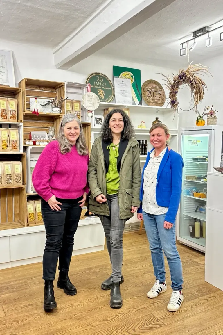Drei Frauen stehen in einem Laden. Die Frau auf der linken Seite trägt einen rosa Pullover. Die Frau in der Mitte trägt eine grüne Jacke. Die Frau auf der rechten Seite trägt eine blaue Jacke. Hinter ihnen befinden sich Regale mit verschiedenen Artikeln.