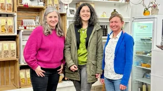 Three women stand in a shop. The woman on the left wears a pink sweater. The middle woman wears a green jacket. The woman on the right wears a blue jacket. Behind them are shelves with various items.