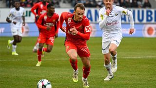 A soccer match is underway on a field, with players in red and white jerseys. One player in red runs towards the ball, while another in white follows closely behind. The player in red looks focused, and the player in white has a determined expression.