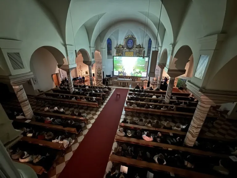 A crowd gathers in a church, watching a film projected on a large screen in front of the altar. The church has arched ceilings and a checkered floor.