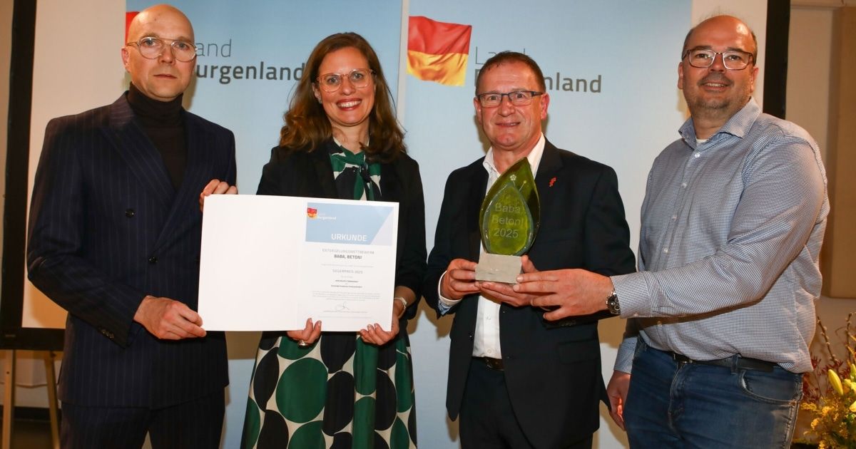 Vier Personen stehen vor einem blauen Hintergrund mit einer deutschen Flagge. Eine Frau hält ein Zertifikat und ein Mann hält einen Pokal mit der Aufschrift 'Baba Beton 2025'.