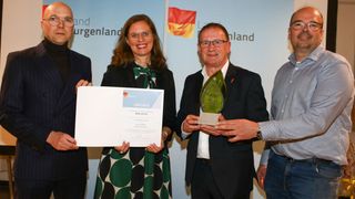 Four people stand in front of a blue backdrop with a German flag. A woman holds a certificate and a man holds a trophy with the text 'Baba Beton 2025'.