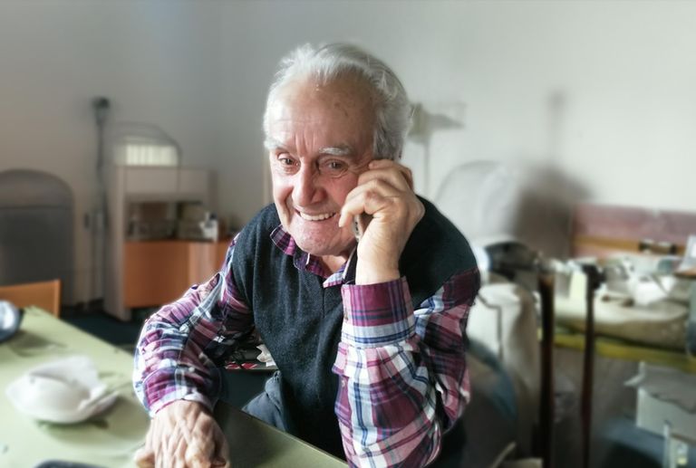 Ein älterer Mann mit weißen Haaren lächelt und telefoniert in einem Raum mit weißen Wänden.