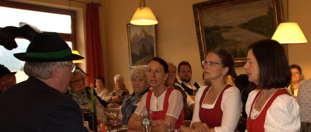 Eine Gruppe von Menschen sitzt in einem Raum, zwei Frauen in roten und weißen Outfits führen ein Gespräch, ein Mann in einem schwarzen Hut spricht mit ihnen, ein Tisch vor ihnen mit Gläsern und Flaschen und ein Gemälde an der Wand.