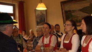 Eine Gruppe von Menschen sitzt in einem Raum, zwei Frauen in roten und weißen Outfits führen ein Gespräch, ein Mann in einem schwarzen Hut spricht mit ihnen, ein Tisch vor ihnen mit Gläsern und Flaschen und ein Gemälde an der Wand.