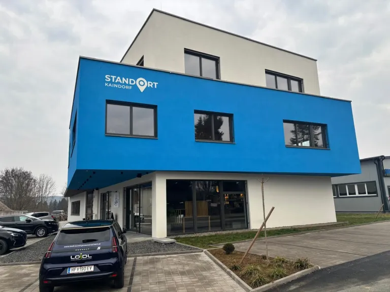 Die Vorderseite eines blauen Gebäudes namens STANDORT KAINDORF, mit großen Glasfenstern, einem geparkten Auto und Grünpflanzen.