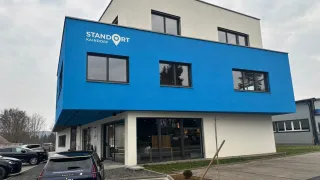 The front of a blue building named STANDORT KAINDORF, with large glass windows, a parked car, and greenery.
