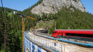 A red train with "railjet" written on it is parked on the track with a yellow crane on the left. Workers are standing on the track. Trees and mountains are behind the track.