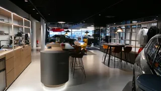 A modern car dealership with several cars on display, featuring a circular reception desk, seating area, and glass shelves. The showroom has white floors, ceiling lamps, and a fire extinguisher.