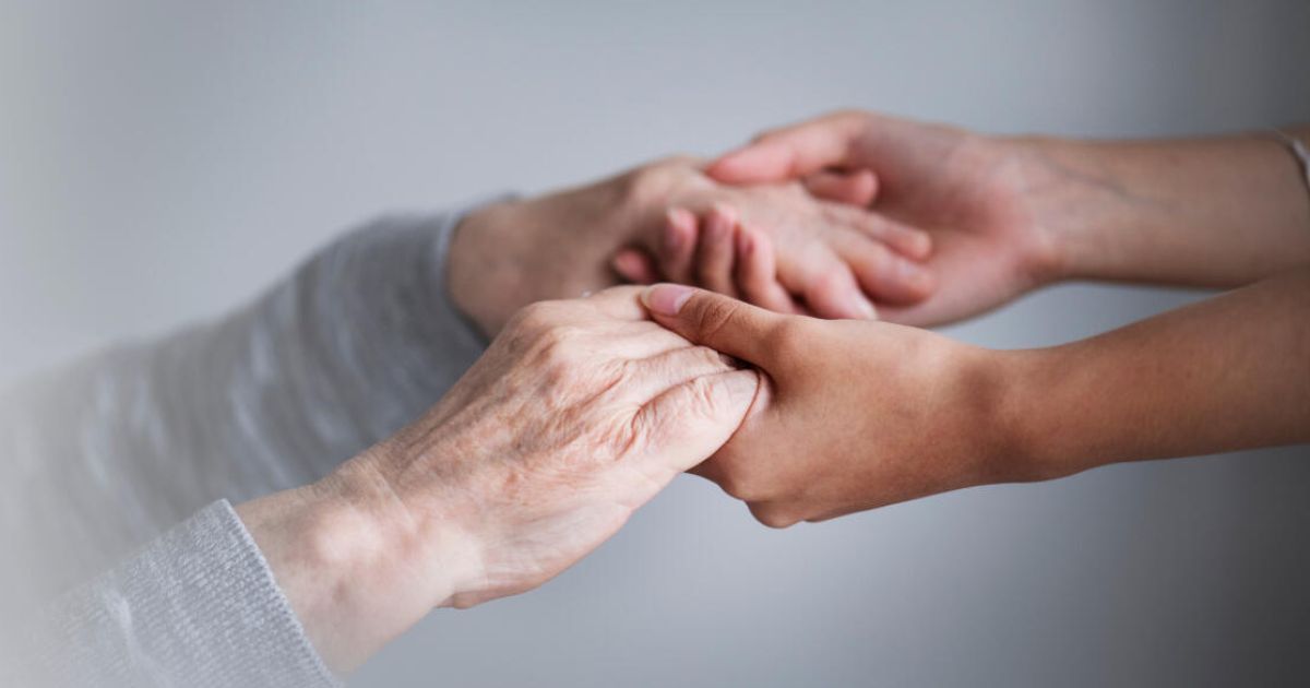 Two hands, one elderly and one younger, are clasped together in a gesture of support and solidarity. The elder hand is wrinkled and has visible veins, while the younger hand is smooth. They are positioned close to each other against a blurred background.