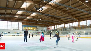 A group of people ice skating in an indoor ice rink. They wear winter clothing and ice skates. The rink has wooden beams and large windows.