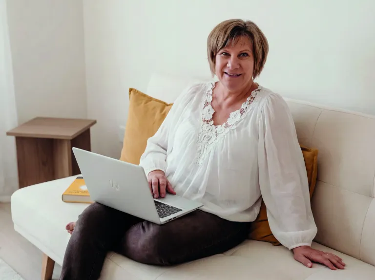A woman with short hair is sitting on a couch, smiling and holding a laptop on her lap. She wears a white blouse and brown pants. A book is on the armrest.